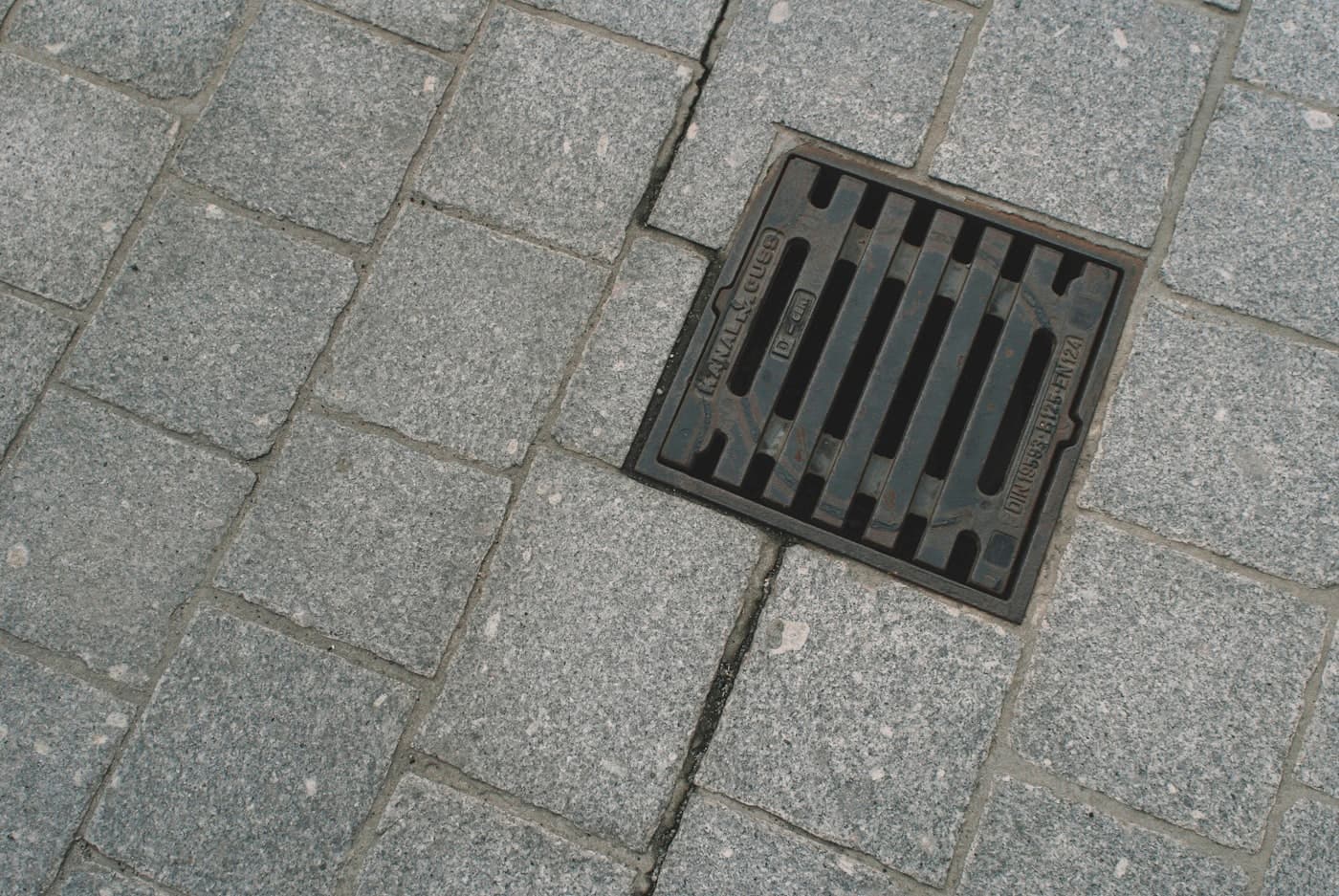 Vierkant gietijzeren putrooster tussen grijze straatstenen.